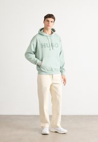 Giovane uomo in felpa verde chiaro "HUGO", pantaloni color crema e sneaker bianchi su uno sfondo bianco semplice.