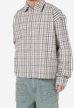 Homme portant une chemise à carreaux beige et blanche boutonnée et un pantalon en jean bleu clair avec poches appliquées cousues.