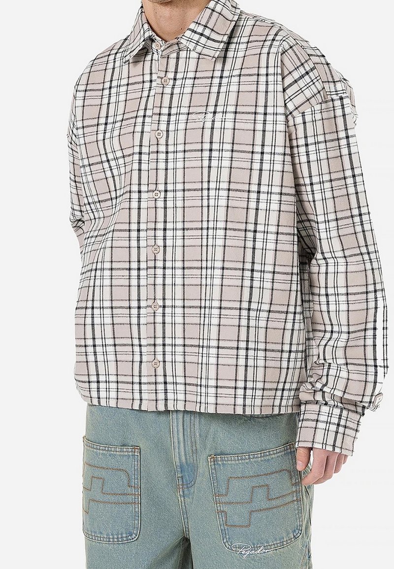 Homme portant une chemise à carreaux beige et blanche boutonnée et un pantalon en jean bleu clair avec poches appliquées cousues.