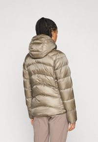Vrouw met gevlochten haar, gekleed in een glanzende taupe pufferjas met een capuchon, staande met haar rug naar de camera tegen een effen lichte achtergrond.