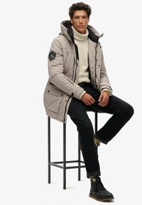 Ljusbeige pufferjacka med huva, med svart foder, sidofickor och en logopatch på ärmen. Matchas med mörka byxor och stövlar.