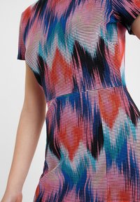 Abito a maniche corte realizzato in tessuto elasticizzato, con un vivace motivo zigzag multicolore in rosso, blu e nero su uno sfondo chiaro.
