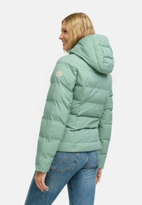 Lichtgroene pufferjack met capuchon, met een doorgestikt ontwerp en een rond logo-opdruk op de linkerarm, gecombineerd met blauwe jeans.