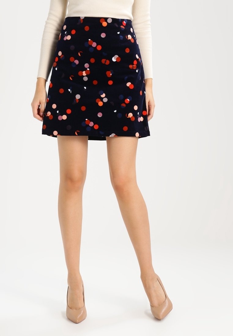 Femme portant une mini-jupe bleu marine à pois multicolores, des talons hauts beiges et un haut à manches longues côtelé crème, debout devant un fond blanc.