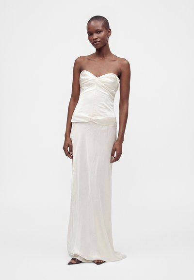 Femme portant une robe bustier en satin écru avec un corsage cintré et une longue jupe, debout devant un fond clair uni.