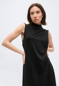 Γυναίκα με κοντά καστανά μαλλιά, ντυμένη με αμάνικο μαύρο φόρεμα ψηλής λαιμόκοψης, στέκεται με τα χέρια στη μέση μπροστά σε απλό φωτεινό φόντο.