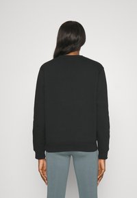 Personne aux longs cheveux foncés portant un sweat-shirt noir uni et un pantalon bleu clair, debout face à un mur blanc, vue de dos.