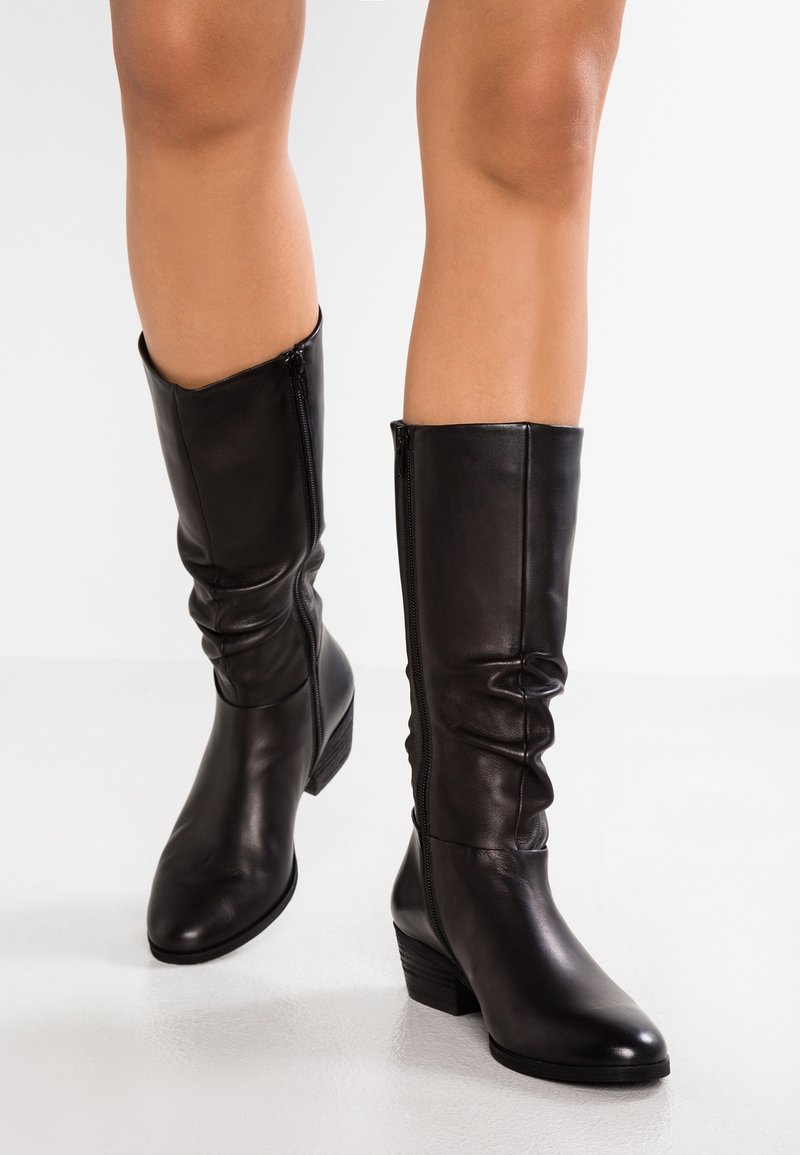 Bottes noires en cuir montant jusqu'au genou, avec une coupe ajustée, un design froissé, des fermetures éclair latérales et un petit talon empilé pour la stabilité. Texture lisse.