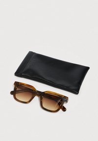 Kilpikonnankuviolliset neliönmuotoiset aurinkolasit ruskeilla liukuvärisillä linsseillä, vieressään musta pehmeä kotelo, jossa on kohopainettu teksti "QUAY", vaalealla pinnalla.