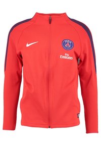 Punainen vetoketjullinen takki, joka on valmistettu sileästä kankaasta. Sen olkapäissä on tummansinisiä yksityiskohtia, Nike-logo ja Pariisin logo, jossa on teksti "Fly Emirates".