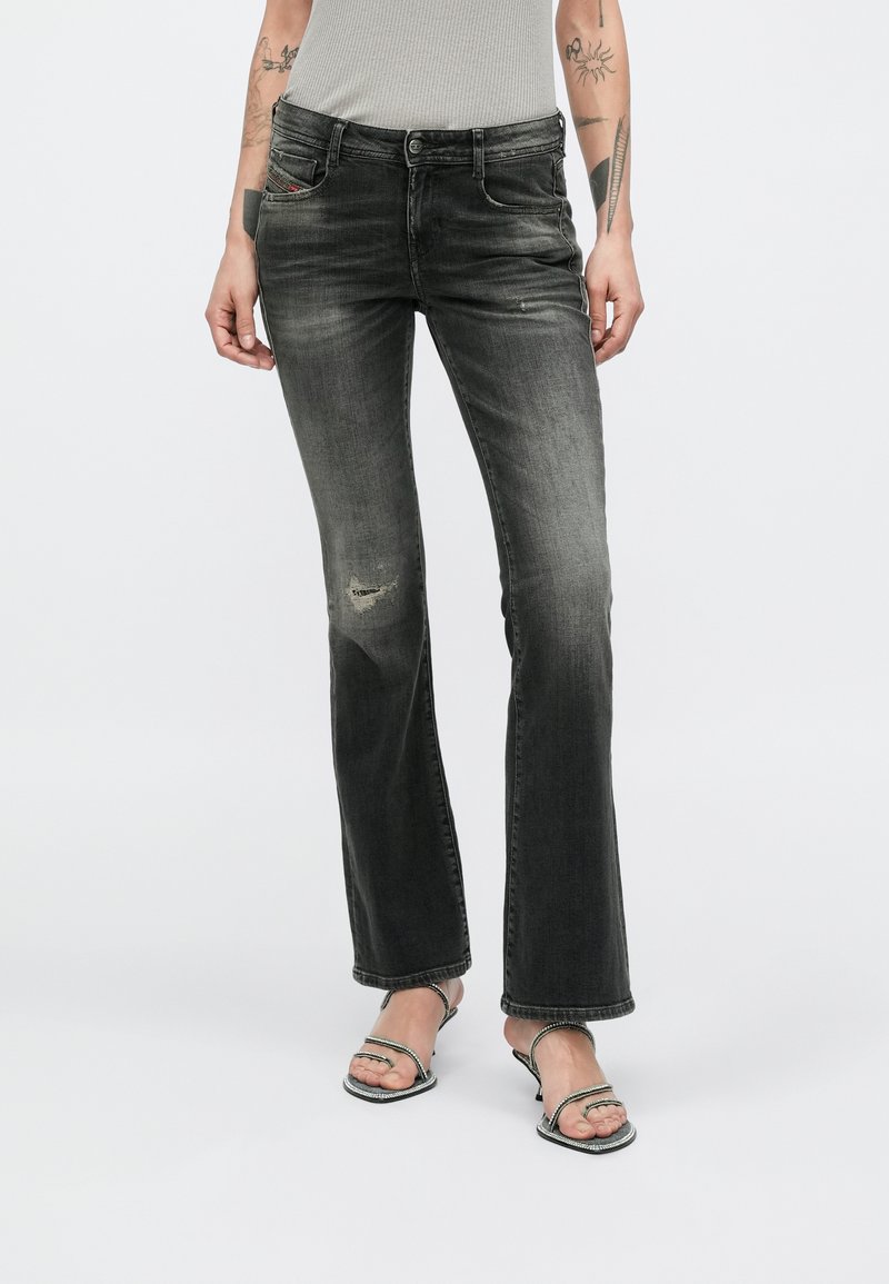 Žena u izblijedjelim crnim trapericama bootcut kroja s blagim oštećenjima na koljenima i srebrnim sandalama na visoku petu s trakicama, stoji na bijeloj pozadini.