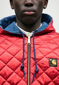 Κουβερτισμένο κόκκινο μπουφάν με κουκούλα επενδεδυμένη με τζιν, ναυτικές κορδόνες και χρυσό φερμουάρ. Διαθέτει μαύρη ετικέτα με το λογότυπο "Carhartt" στο στήθος.