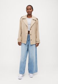 Beige trenchcoat med dobbelspent design, brede ermer og sidelommer, kombinert med lyseblå vide jeans og hvite joggesko.