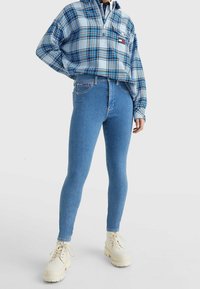 Leggings de mezclilla azul claro con cintura alta, tejido elástico y detalles de costura sutiles; combinados con una camisa de cuadros azul con cremallera y botas color crema.