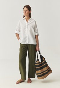 Camicia bianca in lino con bottoni, pantaloni larghi a righe di olive crinkle, e una borsa a tracolla intrecciata a righe nero e marrone. Sandali slide marroni.