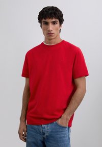 Jonge man met donker krullend haar in een effen rode t-shirt met korte mouwen en blauwe jeans tegen een effen lichtgrijze achtergrond.