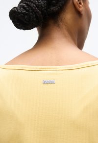 Οπίσθια όψη προσώπου με μαλλιά πλεγμένα σε κότσο που φοράει κίτρινη μπλούζα με μικρή ετικέτα της μάρκας Columbia στο κέντρο κοντά στη λαιμόκοψη.