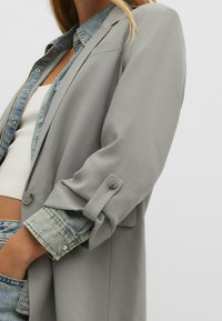 Femme portant un blazer gris par-dessus une veste en jean et un haut court blanc, la main dans la poche de son jean en denim, sur fond uni.