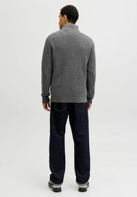 Vīrietis, kas skatās prom, ģērbies pelēkā ribotā augstajā kaklā, tumšās brīvajās džinsēs un pelēkās kedās uz vienkārša balta fona.