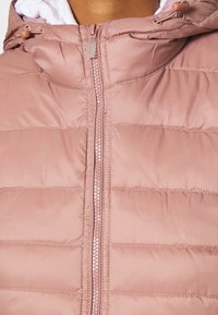 Veste en duvet matelassée rose clair avec un col montant, fermeture éclair complète et capuche avec cordon de serrage. Tissu lisse et détails de couture subtils.