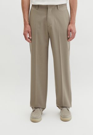 Mann trägt beige weite Hosen und passende hellbeige Wildlederschuhe vor einem schlichten hellen Hintergrund.