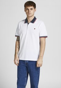 Balta polo krekls ar tumši zilu apkakli, sarkaniem un tumši ziliem svītrām uz piedurknēm, trīs pogu piegriezums un neliels dekoratīvs emblēma krūtīs.