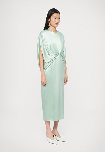 Φόρεμα από σατέν σε μέντα πράσινο χρώμα με λεπτομέρεια στρίφωμα στη μέση, κοντά μανίκια και ίσια σιλουέτα. Το μοντέλο συνδυάζεται με λευκά παπούτσια με τακούνι.