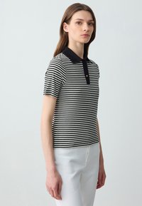 Polo a righe in bianco e nero con colletto nero uniforme, maniche corte e patta con bottoni; realizzato in un tessuto a maglia testurizzato.