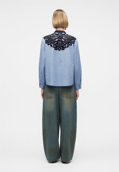 Άτομο με κοντά ξανθά μαλλιά που φοράει μπλούζα με μπλε ρίγες και μαύρη δαντέλα στην πάνω πλάτη, καθώς και φαρδιά ξεβαμμένα τζιν παντελόνια.