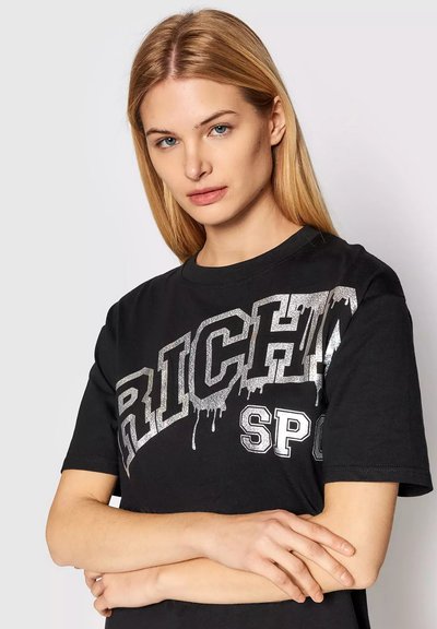 Donna bionda con occhi azzurri che indossa una maglietta nera con il testo "RICH" in argento colante e le braccia incrociate su uno sfondo semplice.