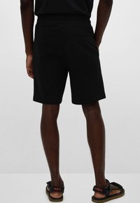 Sorte shorts med en glatt tekstur, laget av lett stoff. Har en baklomme og er paret med sorte sandaler som har merkevaren.