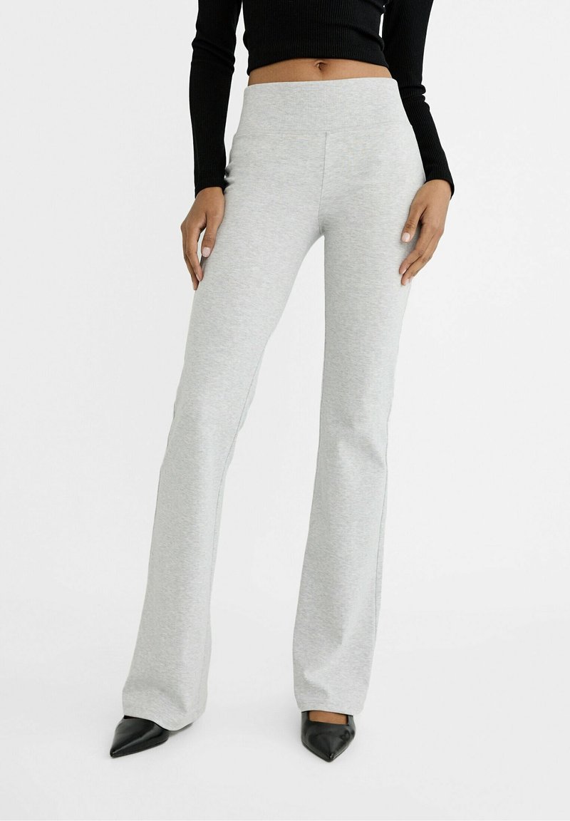 Femme portant un pantalon évasé taille haute gris clair, des chaussures noires à bout pointu et un top noir court à manches longues, debout devant un fond blanc.