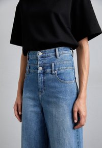 Personne portant un jean bleu taille haute avec fermeture à double bouton et un haut noir à manches courtes, debout devant un fond uni.