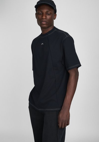 T-shirt noir en coton à manches courtes, avec des accents de couture blancs et un petit logo sur le devant, associé à un jean foncé.