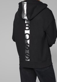 Svart huvtröja med en glänsande silver vertikal "BOSS" logotyp på baksidan, med mjuk textur och lös passform.