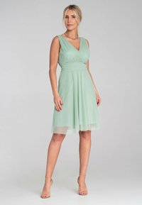Ein mintgrünes Kleid mit einem taillierten Oberteil und einem ausgestellten Rock, das über einen V-Ausschnitt und Schichten aus leichtem Material verfügt. Transparente Tüllakzente.