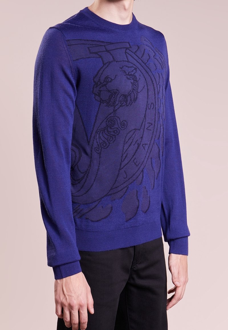 Pull bleu royal en mailles douces. Présente un design graphique sombre d'un lion et un texte stylisé. Manches longues avec des poignets côtelés.