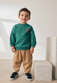 Hanorac verde din bumbac cu imprimeu floral roșu, asortat cu pantaloni în carouri maro și crem. Tenisi negri cu benzi Velcro completează ținuta.