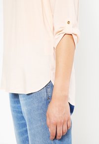 Kvinna som bär en ljus persikofärgad blus med uppvikt ärm och detaljer i guldknappar, samt blå denim jeans som är avskurna vid höften och ärmen.