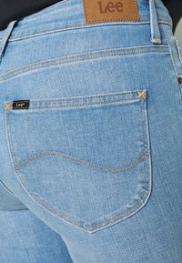 Jeans de mezclilla azul claro con una textura desteñida. Cuenta con un parche de piel en la parte posterior y dos bolsillos traseros con costuras amarillas contrastantes.