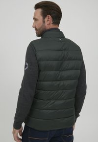 Dunkelgrüne Herren-Pufferweste mit hohem Kragen und gerippten grauen Ärmeln, mit horizontalen Nähten und einem Logo-Patch am Arm.