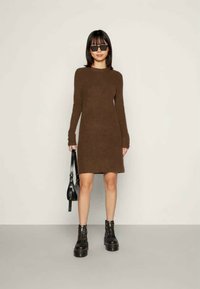Καφέ πλεκτό φόρεμα φούτερ με μακριά μανίκια, εφαρμοστή σιλουέτα και ραβδωτή υφή. Φορεμένο με μαύρες πλατφόρμες και μια μικρή μαύρη τσάντα.