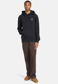 Felpa nera con tasca frontale e coulisse, con un piccolo logo verde; abbinata a pantaloni marroni e scarpe marroni.