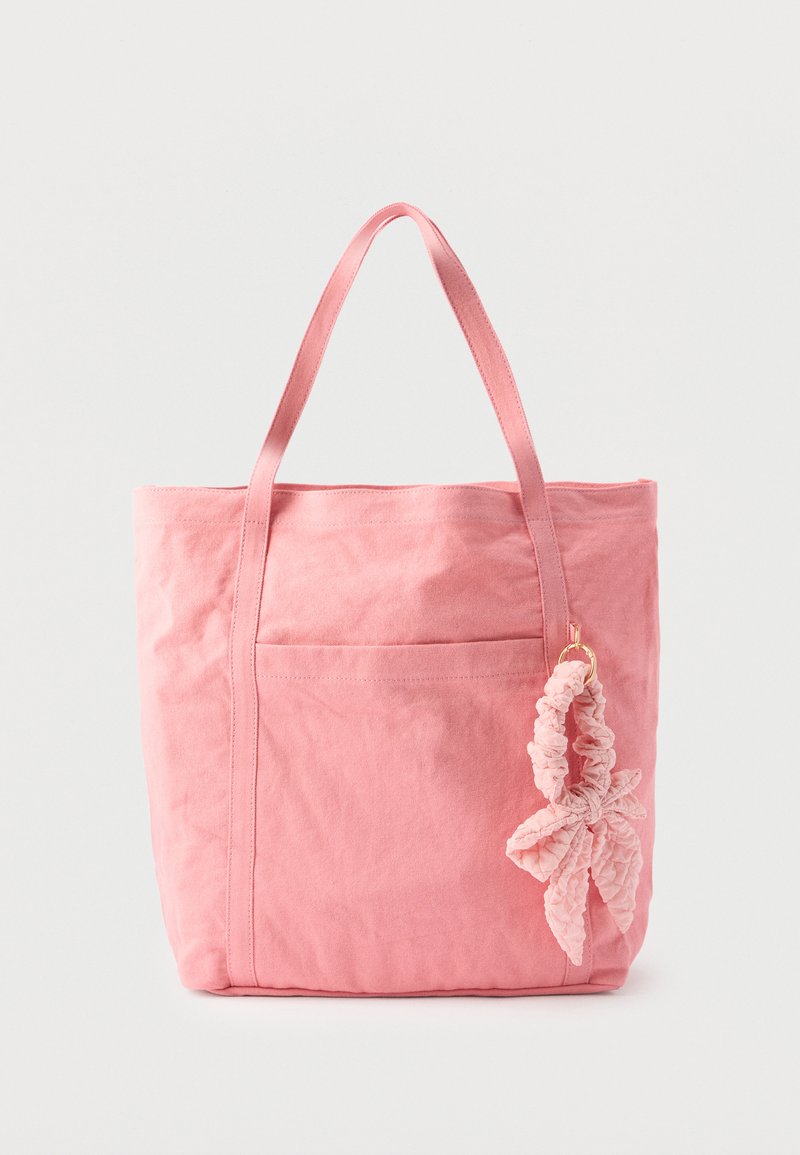 Sac fourre-tout en toile rose avec deux anses et un scrunchie en tissu rose attaché en forme de feuille sur fond blanc.