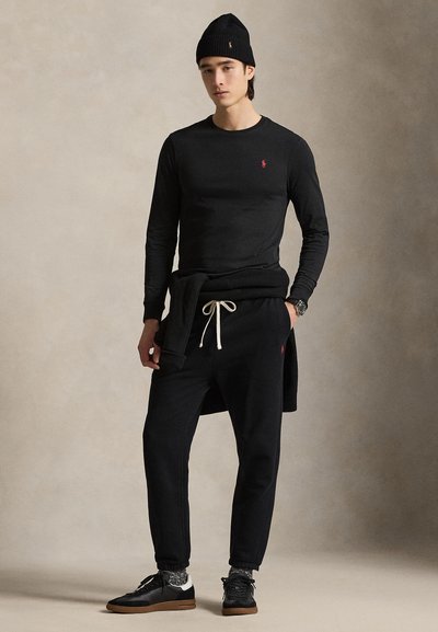 Młody mężczyzna w czarnej czapce, koszuli z długim rękawem, spodniach dresowych, trampkach i swetrze związanym wokół talii, stojący na neutralnym tle.