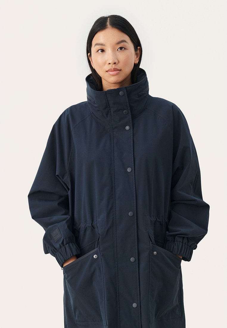 Giacca impermeabile blu navy con colletto alto, polsini elasticizzati e due tasche frontali. Presenta bottoni a pressione e un cordino in vita per una vestibilità regolabile.