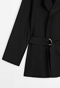 Veste noire à manches longues avec une ceinture en tissu passant à travers une boucle argentée, montrant la poche latérale et une partie du col.