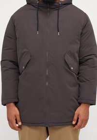 Mörkgrå vadderad jacka med huva, dragkedjestängning och två sidofickor. Har elastiska ärmslut och en slät yta.