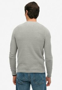 Mężczyzna o ciemnych włosach, ubrany w jasnoszary, teksturowany sweter z długimi rękawami i niebieskie dżinsy, odwrócony plecami do białego tła.