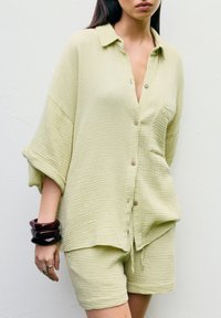 Lichtgroene, gestructureerde linnen blouse met knopen en een zak, gecombineerd met bijpassende shorts. Het model draagt gestapelde donkere bangle armbanden.
